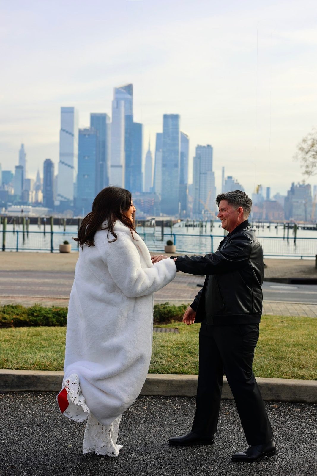 Ashley and Eli holding hands on a waterfront path with the Manhattan skyline behind them