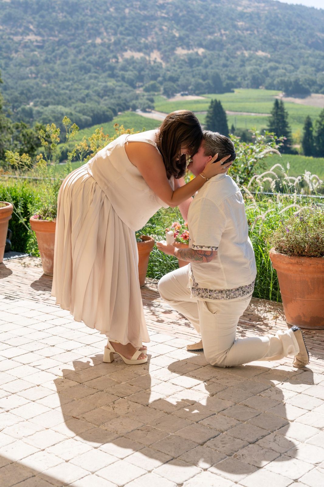 Ashley and Eli's proposal on a vineyard hillside, Ashley kneeling to hold Eli's face while Eli kneels on one knee