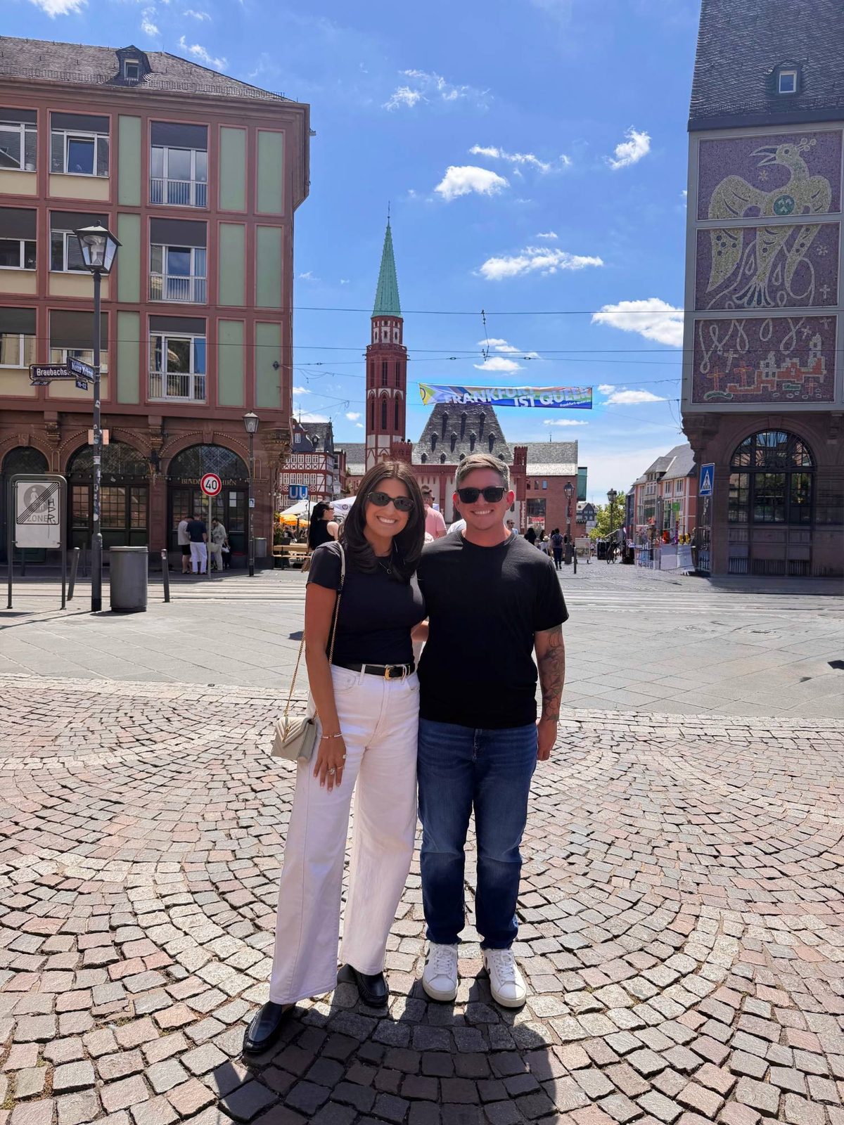 Ashley and Eli in a town square in Frankfurt, with a rainbow Pride banner overhead