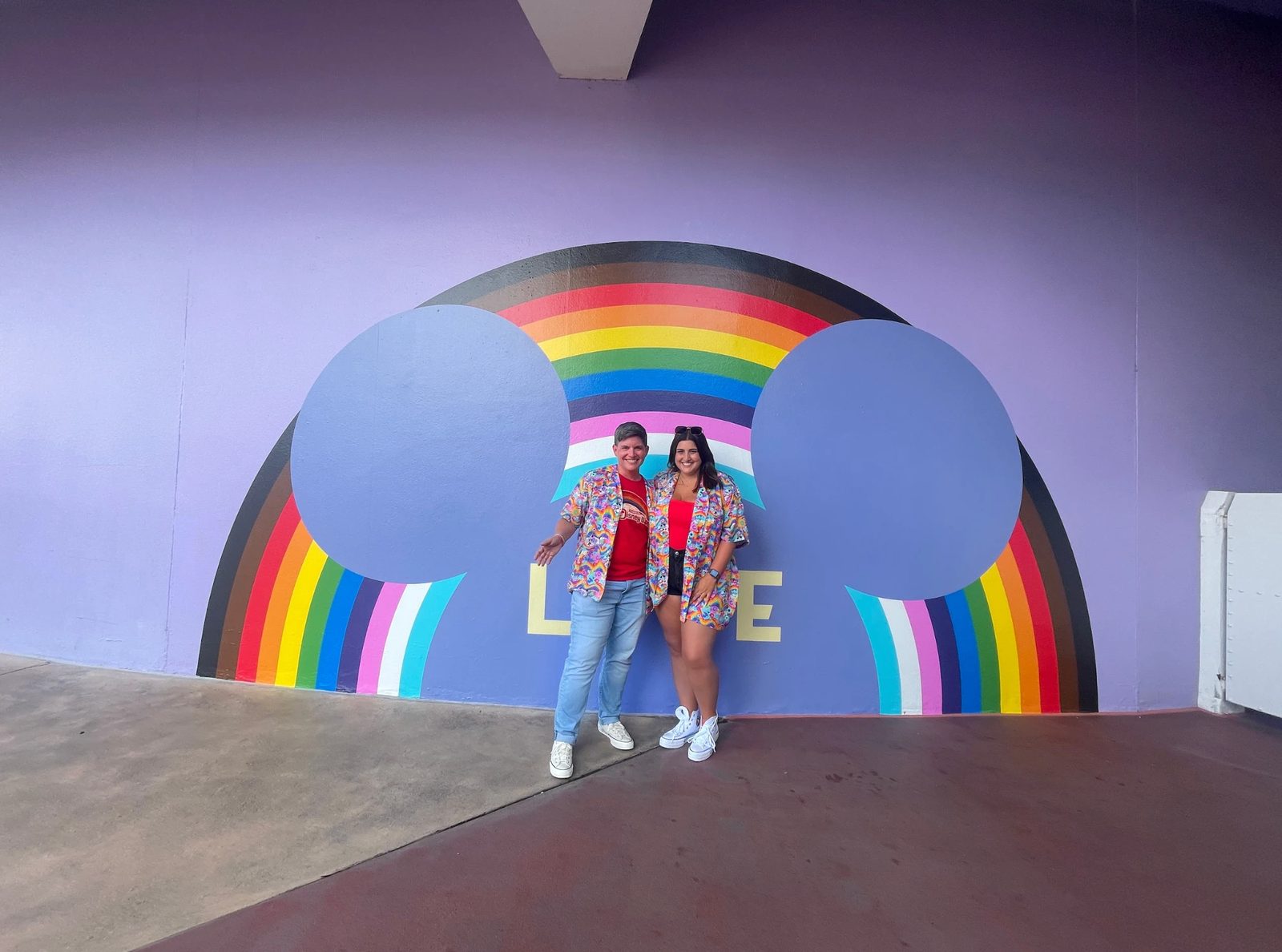 Ashley and Eli in matching shirts in front of a rainbow 'Love' mural