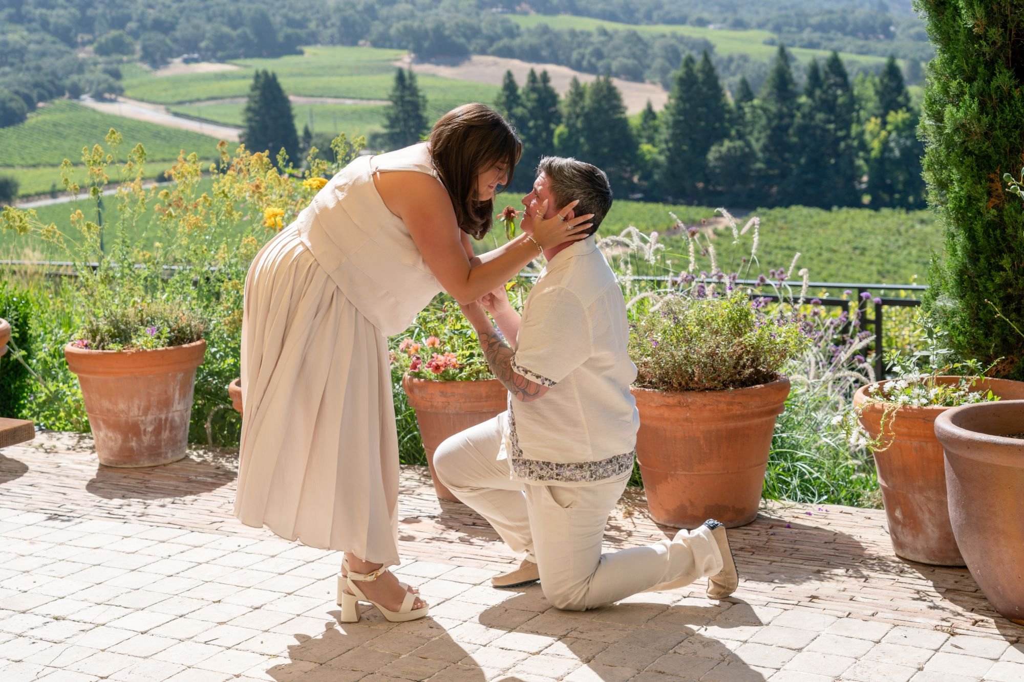 Ashley and Eli's proposal on a vineyard hillside, Ashley kneeling to hold Eli's face while Eli kneels on one knee
