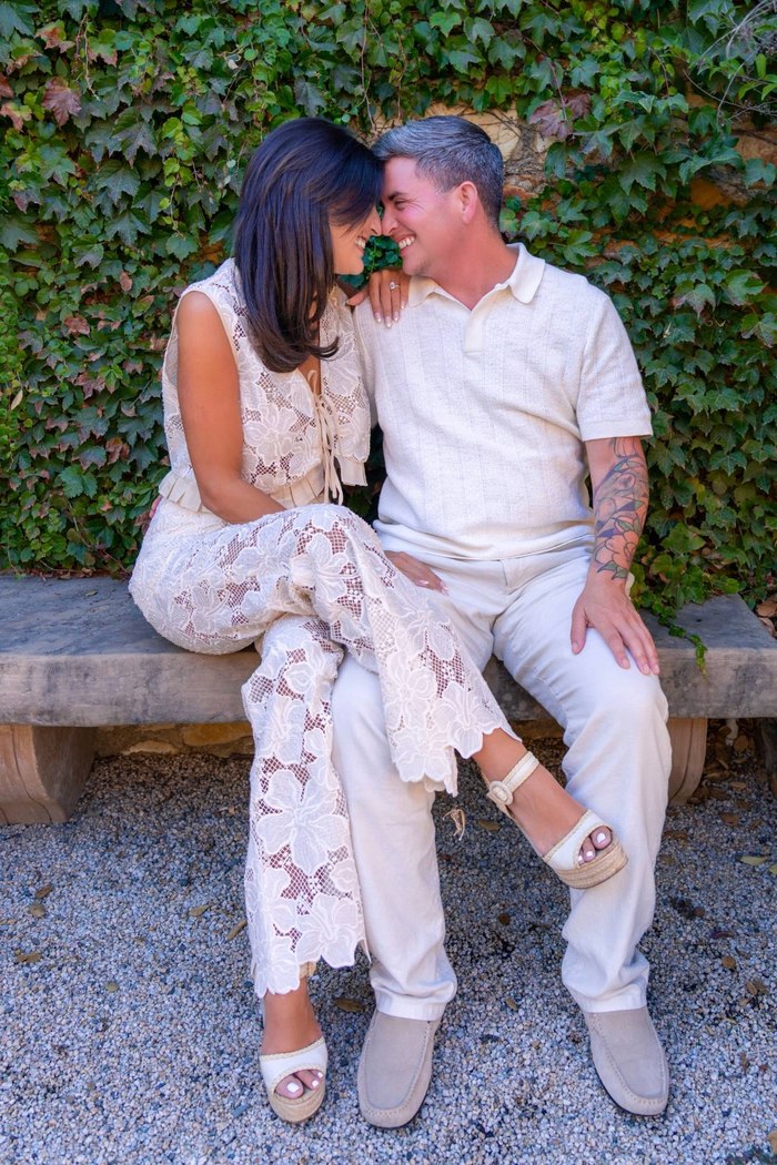 Ashley and Eli sit together on a stone bench, smiling and touching foreheads in front of an ivy-covered wall.